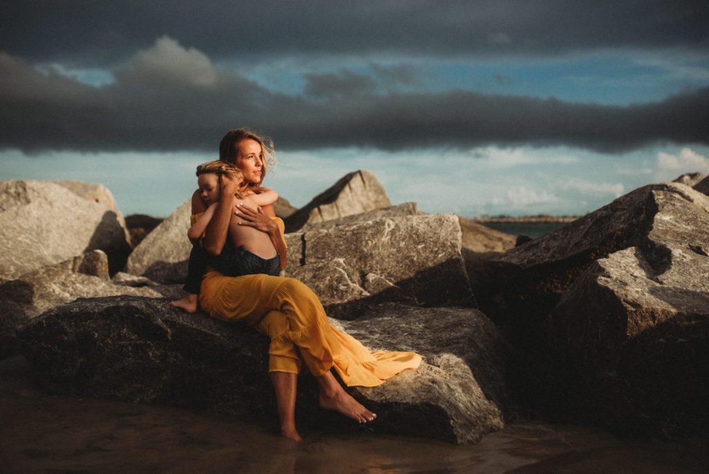 Mother in a yellow dress lovingly embraces her child while sitting on rocky shore, capturing a serene moment