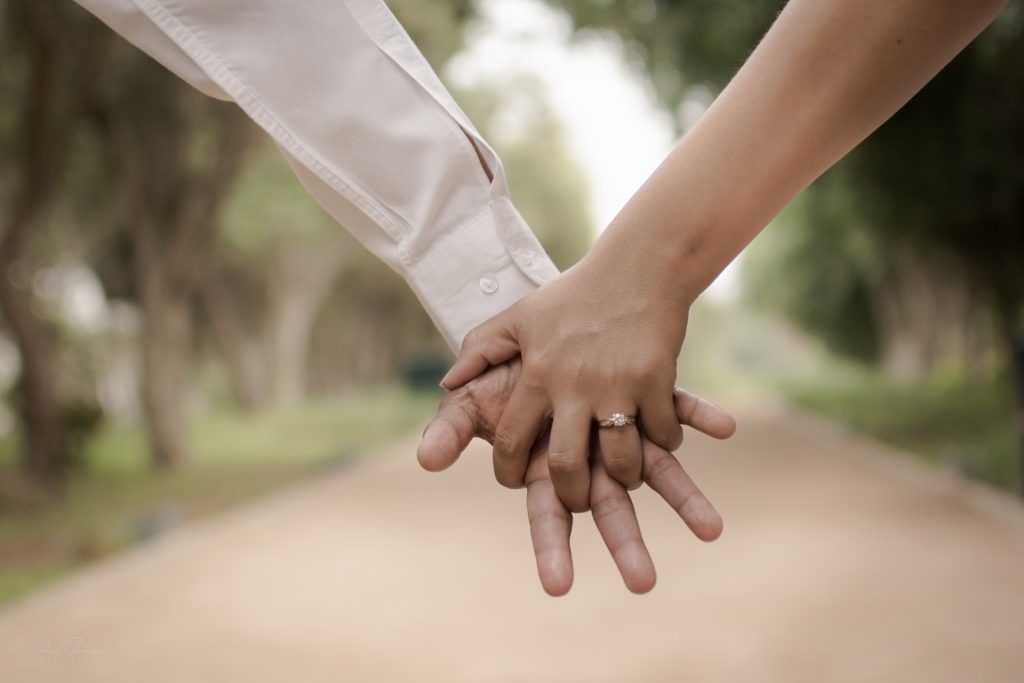 Couple holding hands with a visible engagement ring, walking down a tree-lined path