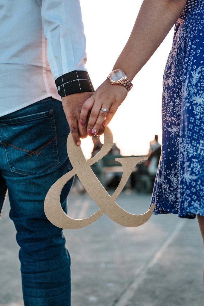 Couple holding hands with an ampersand symbol between them, highlighting their engagement rings 