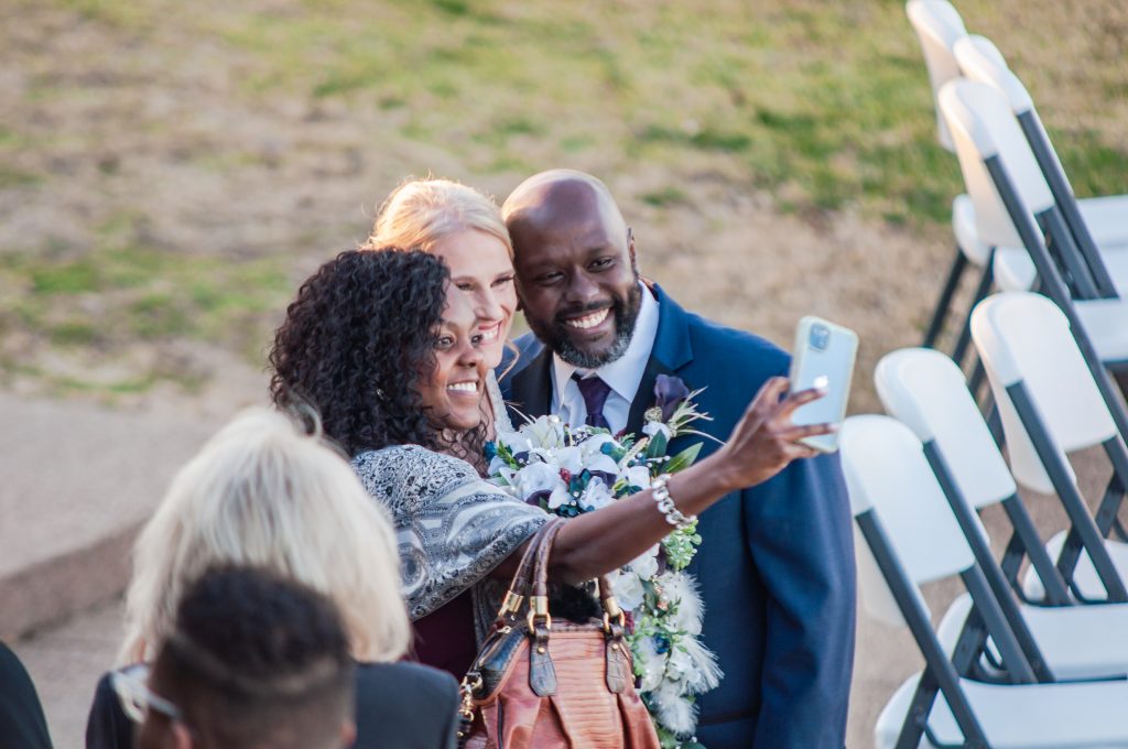 Happy couple taking a selfie with a wedding guest outdoors, capturing a joyful and candid moment during the celebration