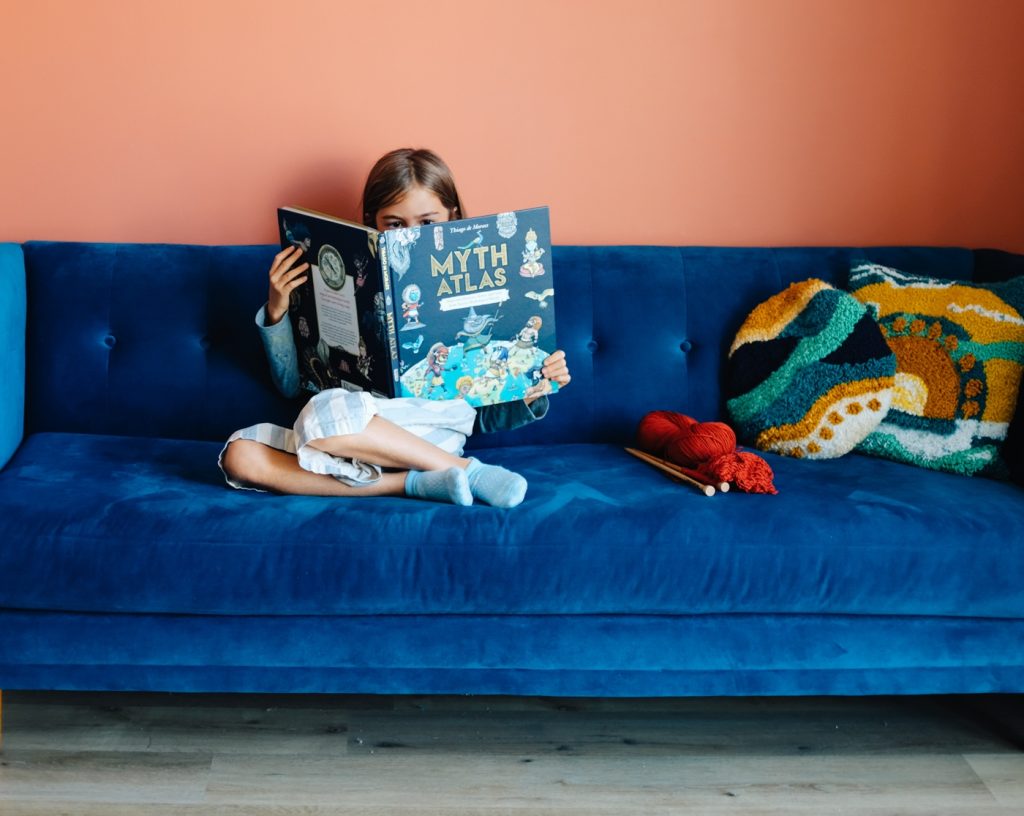 Girl reads ‘myth atlas’ book on blue velvet couch with knit throw pillows