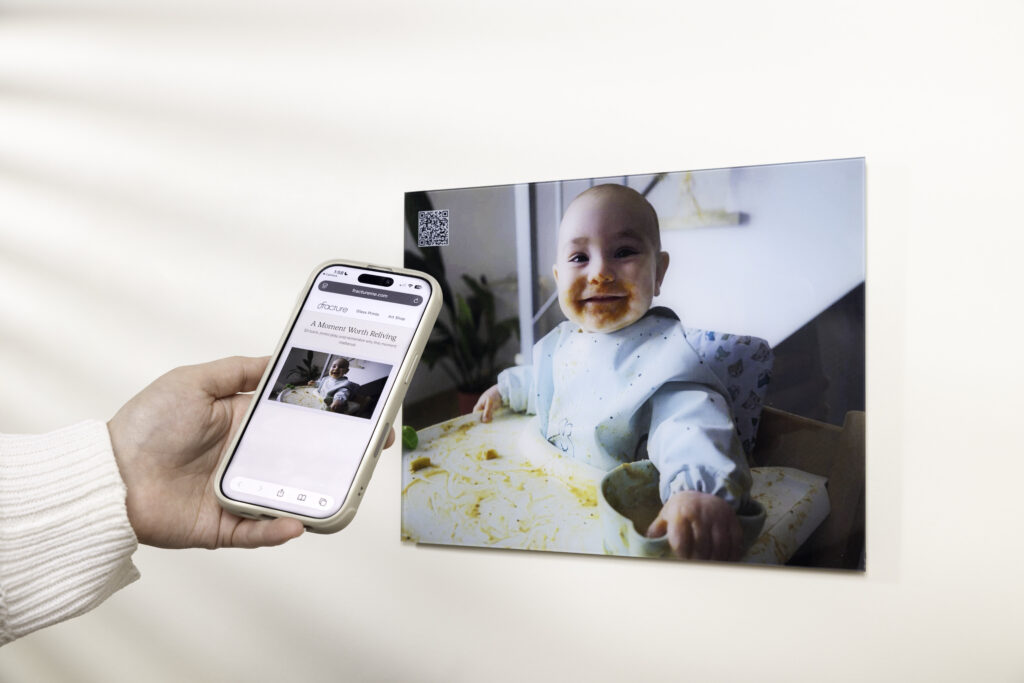 A photo of a cute baby smiling with spaghetti sauce on his face in his high chair. A hand holds a phone, having just scanned a qr code to look back at the video memory connected with the print.