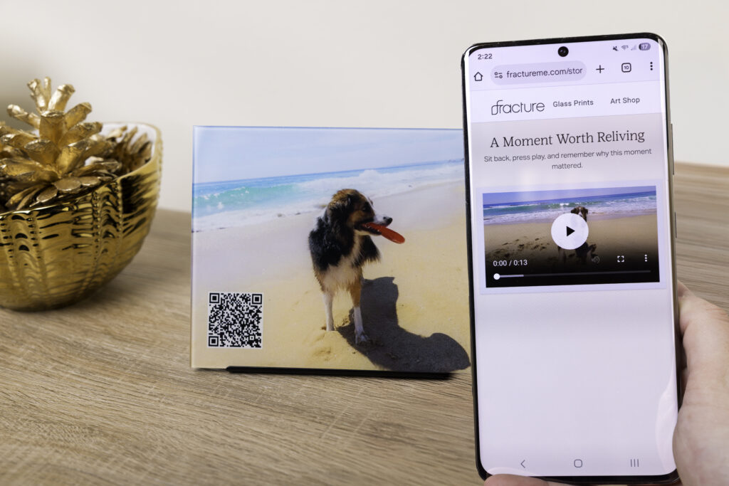 A phone is held in front of a glass print, scanning a QR code that links to a landing page where your video is ready to play and relive any time.