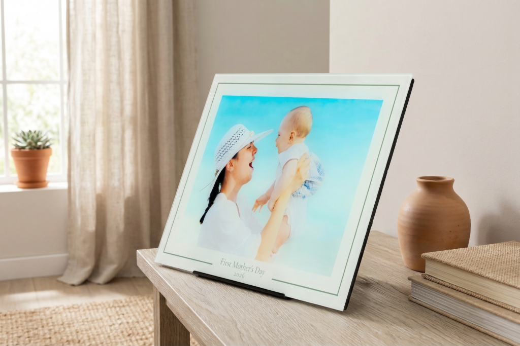 A First Mother's Day gift glass print—with an elegant border that says First Mother's Day 2026—features a photo of a mom lifting her baby in the air. The glass prints its in a stand on a credenza in a room with neutral decor.