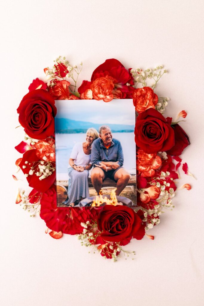 A glass anniversary gift—a glass print of an older couple sitting in front of a fire pit with a beautiful lake behind them—sits atop an arrangement of roses and other flowers.